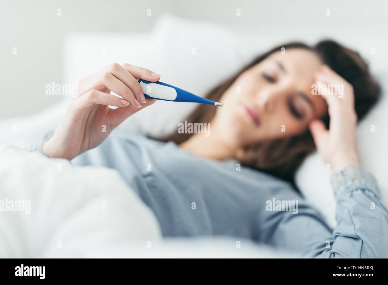 Woman with flu virus lying in bed, she is measuring her temperature with a thermometer and touching her forehead Stock Photo