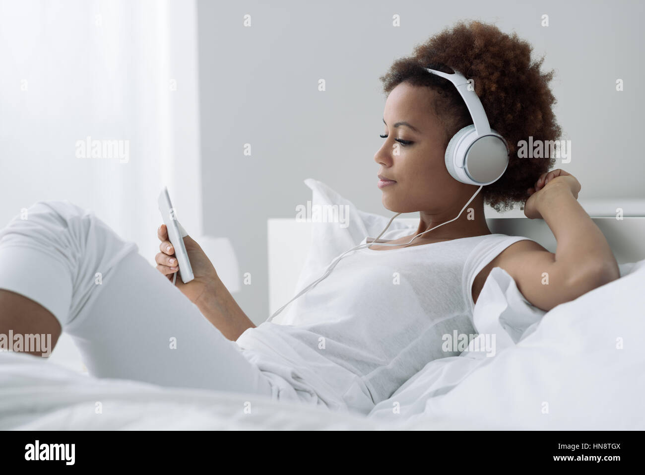 Young african american woman relaxing on the bed and connecting with a touch screen tablet, she is smiling and wearing headphones Stock Photo