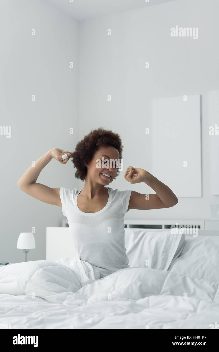 Young african woman waking up early in the morning, she is sitting in her bed and stretching Stock Photo