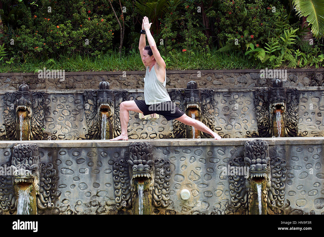 Middle-Aged Asian Man Holds a Hatha Warrior Yoga Pose at Banjar Hot Springs Pools in Bali, Indonesia as Water Pours from Dragon-Like Creatures' Mouths Stock Photo