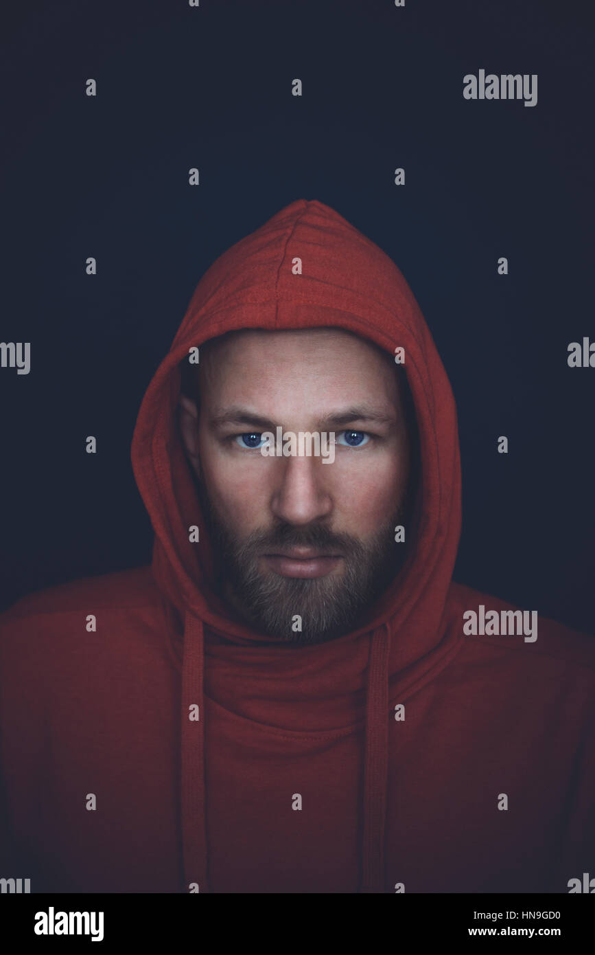 Portrait of a caucasian man in mid thirties, shot in studio wearing a red hooded sweatshirt on a dark blue wall. Stock Photo