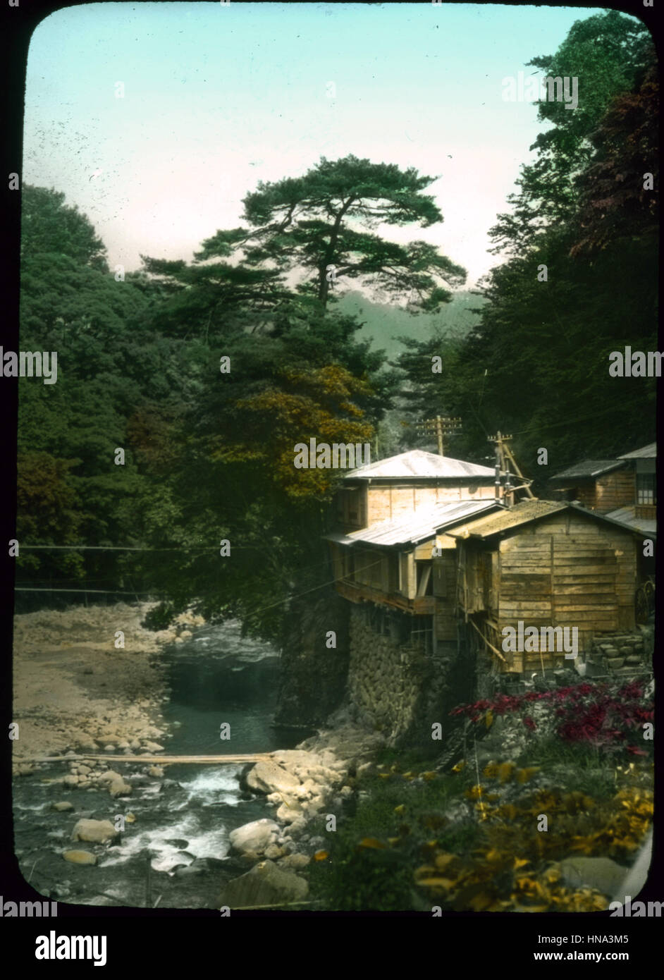 Herbert Geddes, Life in Japan, ca. 1910 -  Buildings built up on stone walled riverbank with trees in background Stock Photo