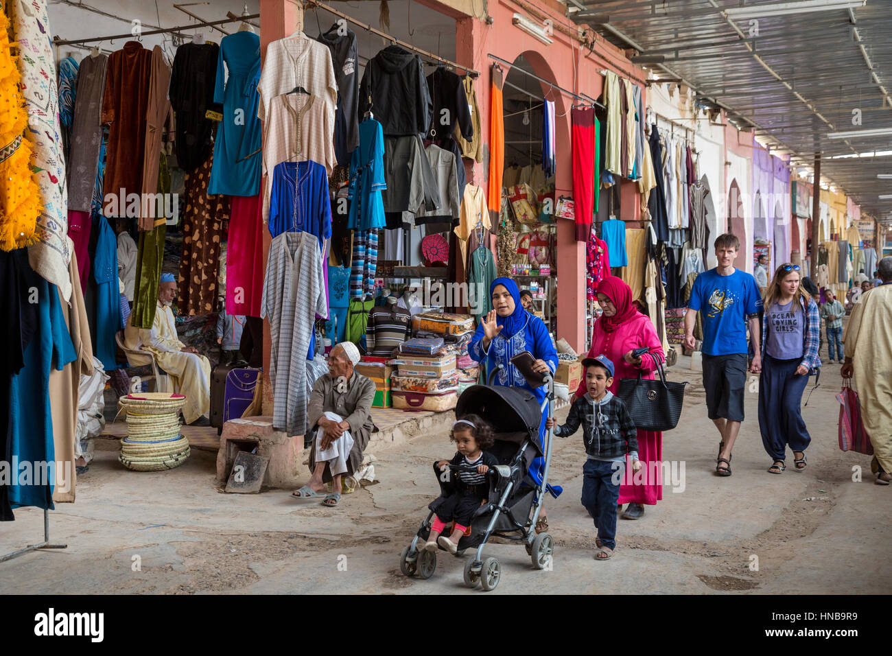 Rissani, Morocco.  Women, Children, and Tourists Walking Past Clothing Shops in the Market. Stock Photo