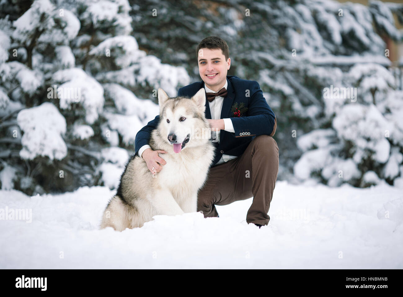 Groom in winter with dog malamute sitting in snow. He hugging dog and