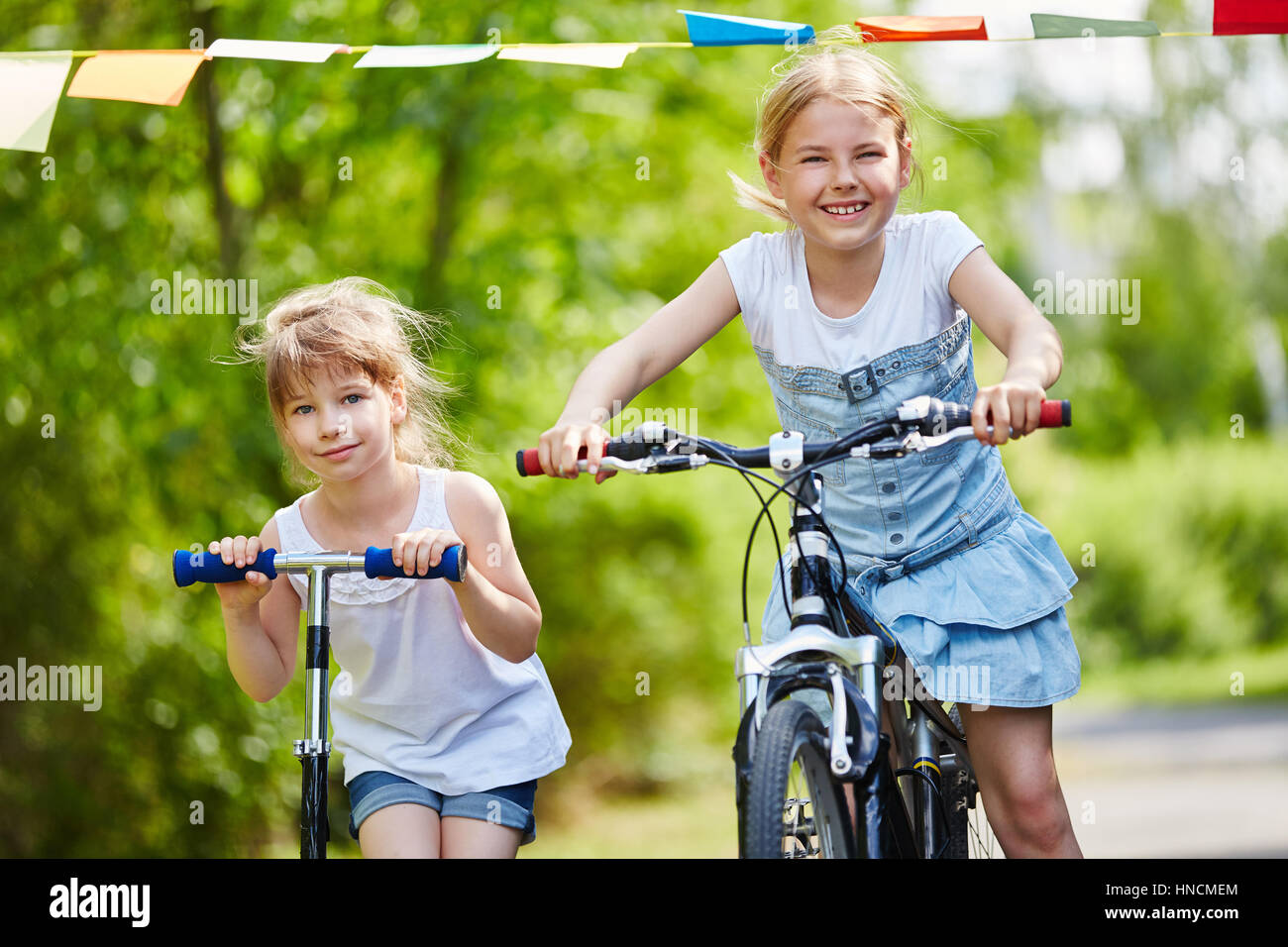Two girls as sisters driving with bicycle and scooter in the park Stock