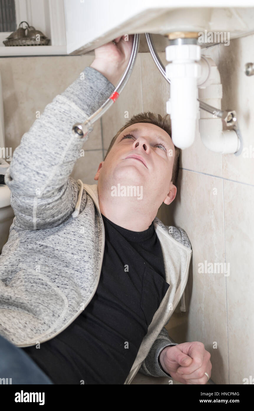 Young man attempting Do It Yourself (DIY) plumbing at home under a sink fitting a new tap looking upwards. Stock Photo