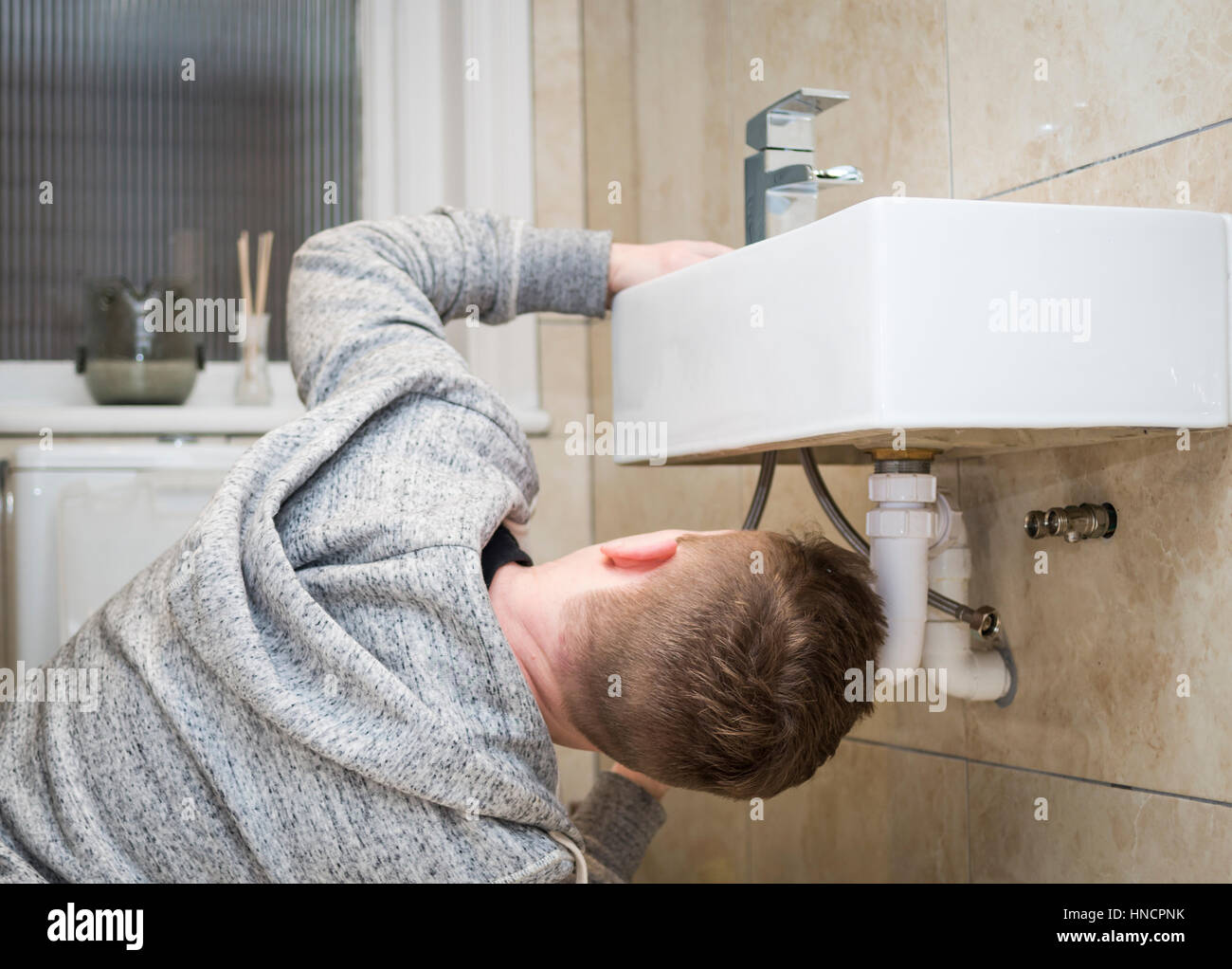 Rear view of a young man attempting Do It Yourself (DIY) plumbing at home under a sink fitting a new tap. Stock Photo