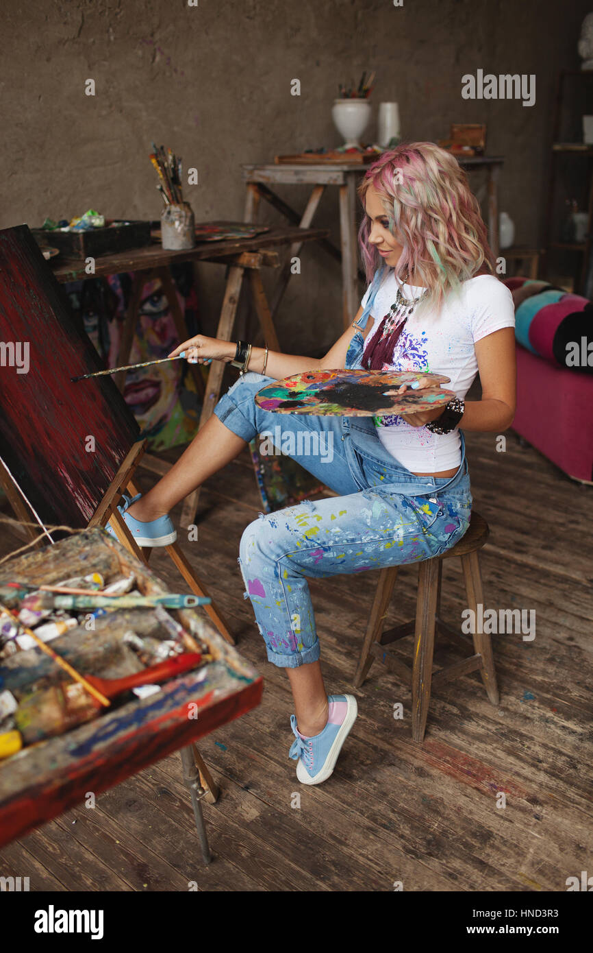 The artist with multi-colored hair in paint-stained jeans paints Stock Photo