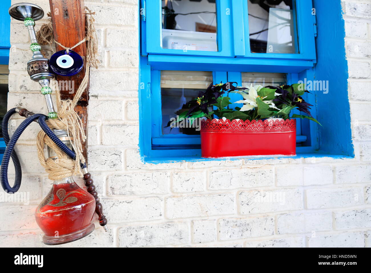 Old hookah-shisha-narghile-waterpipe with nazar-blue bead amulet hanging on the whitewashed wall of a house with blue window and red plantpot-street o Stock Photo