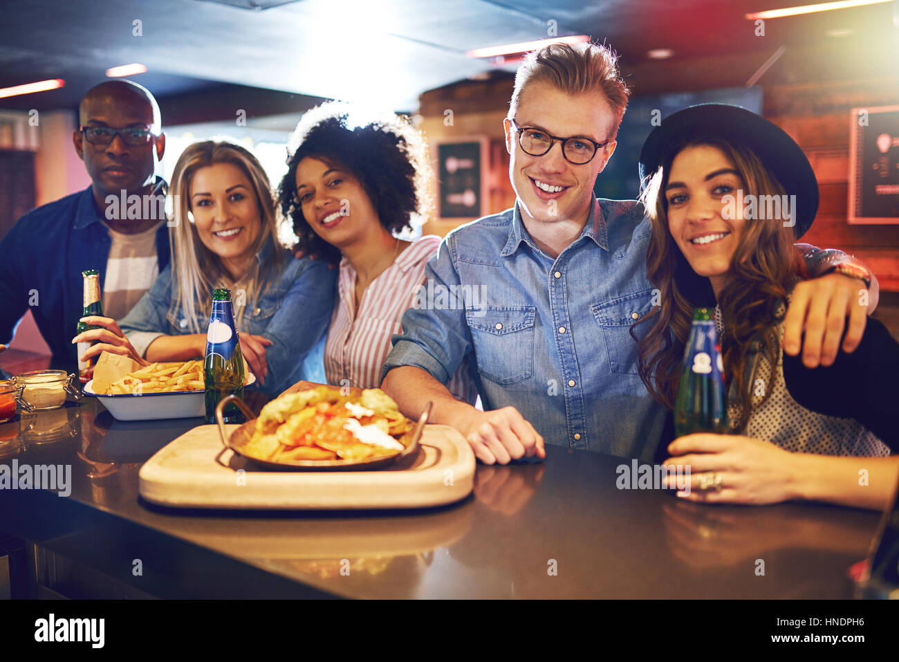 Cheerful friendly people posing at the bar counter with beer and snacks. Horizontal indoors shot. Stock Photo