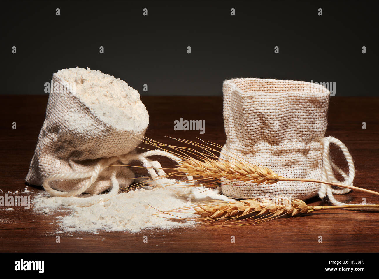 Burlap bag of flour, empty burlap bag and wheat ears on dark wooden table on grey background Stock Photo