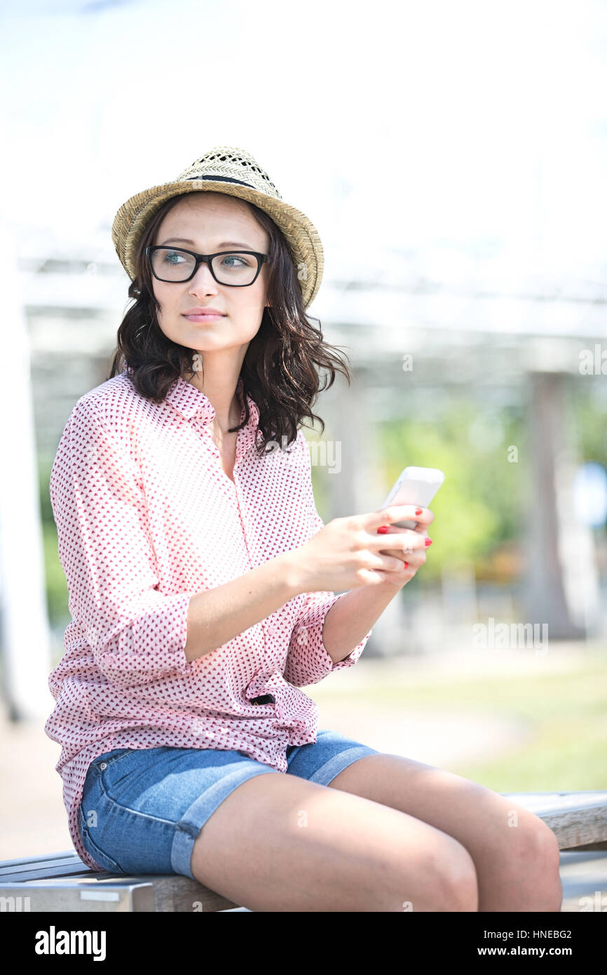Woman looking away while holding mobile phone on bench outdoors Stock Photo