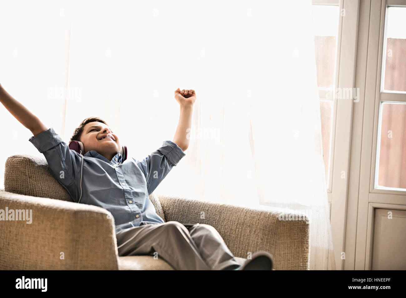 Happy boy with arms raised enjoying music while relaxing on armchair at home Stock Photo
