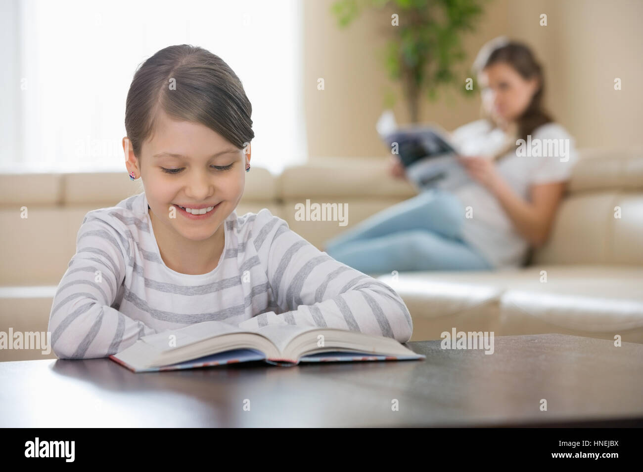 Smiling girl reading book with mother in background at home Stock Photo ...