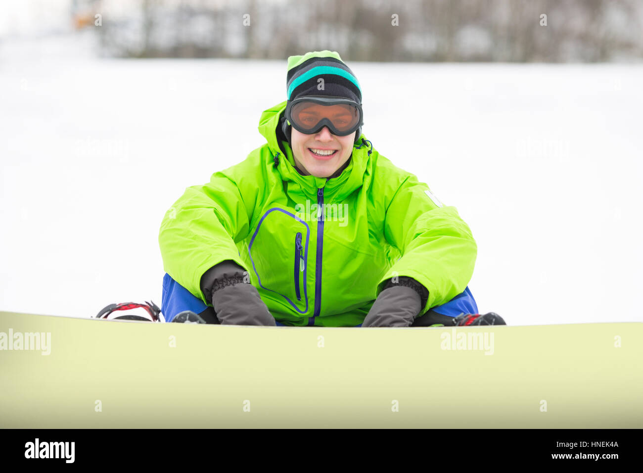 Portrait of smiling young man with snowboard sitting on snowy land Stock Photo