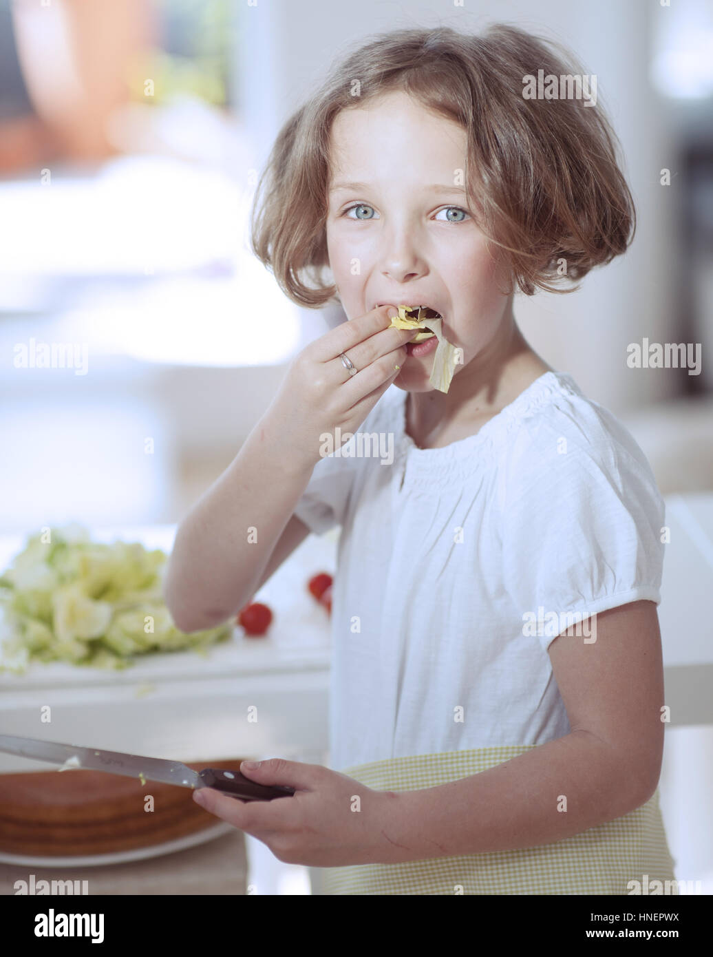 Young girl eating salad whilst holding knife in kitchen Stock Photo