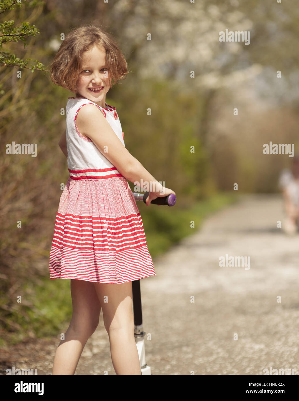 Young girl riding scooter in park away from camera to mother Stock Photo