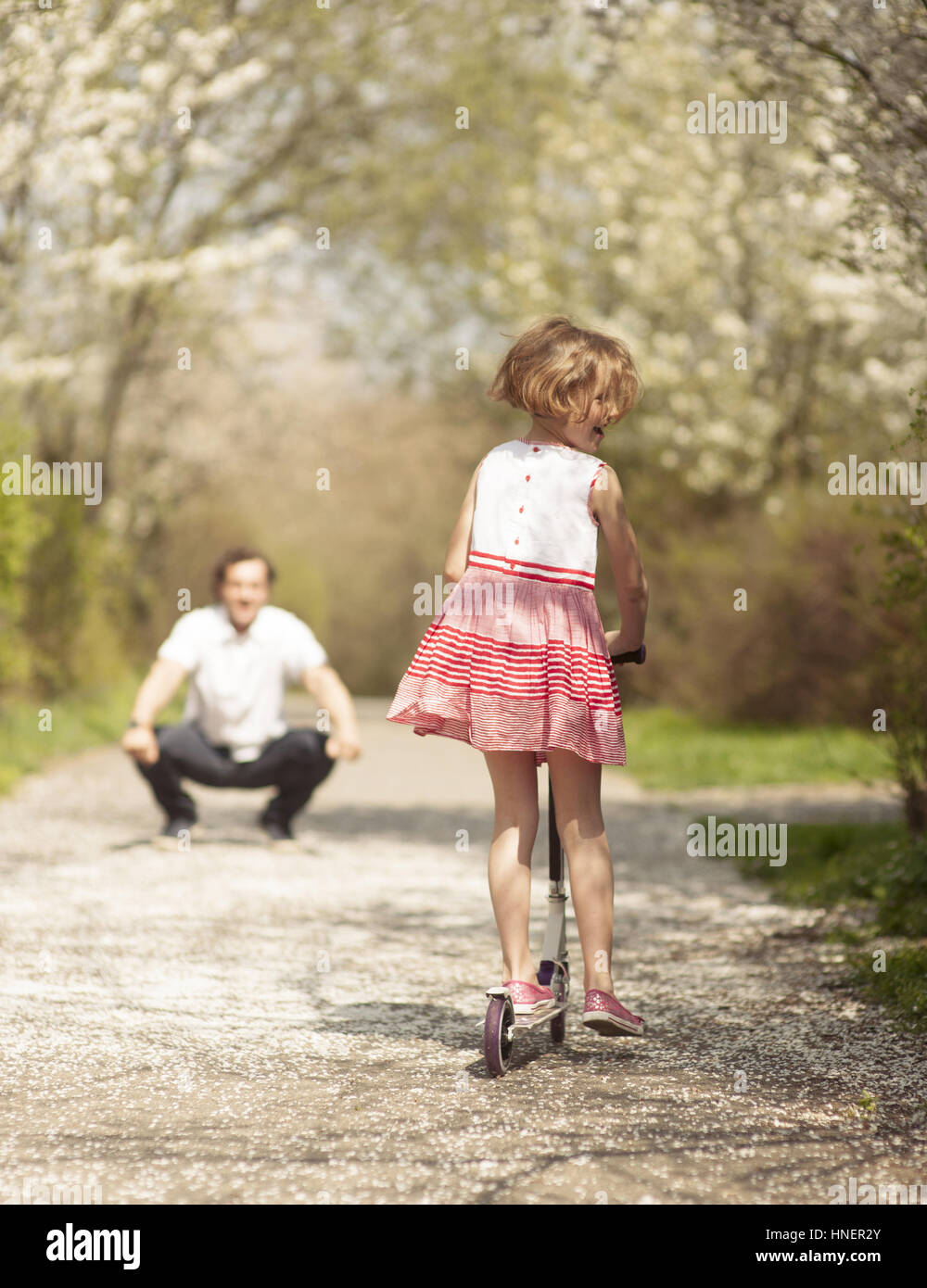 Young girl riding scooter away from camera to father in park Stock Photo