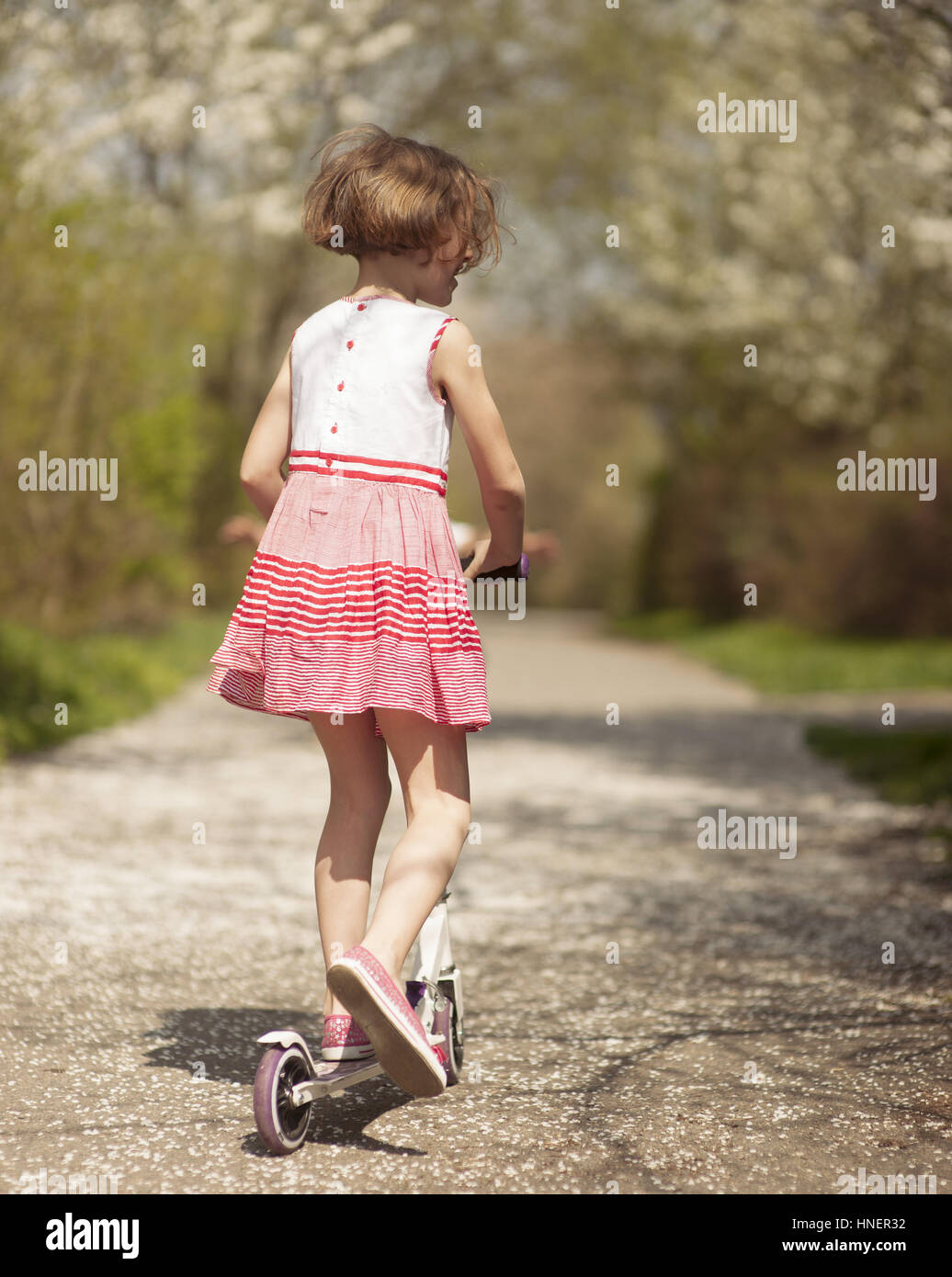 Young girl riding scooter away from camera on path through park Stock Photo