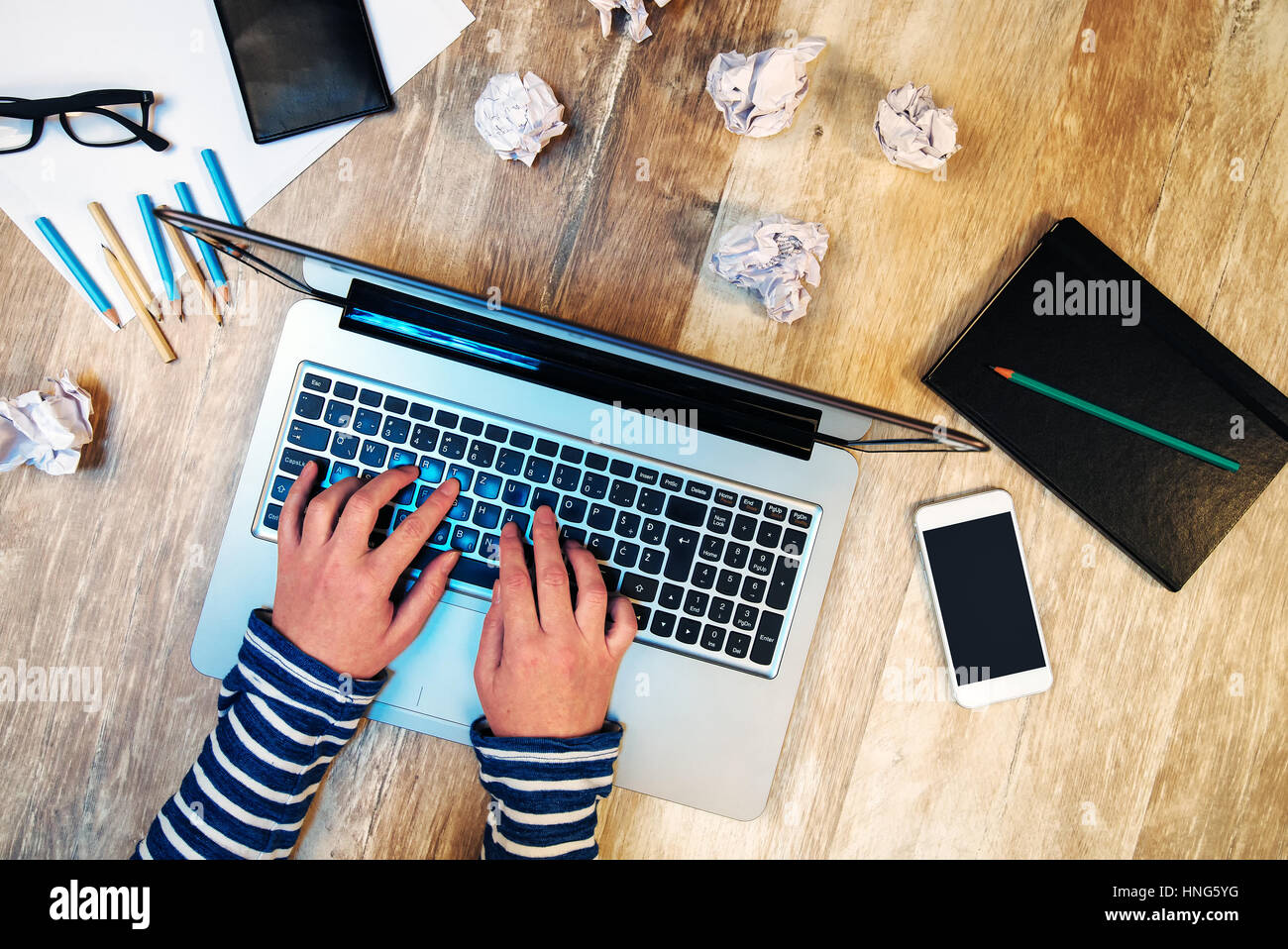 Creative female person working on laptop computer in the office, top view of hands typing keyboard on desk with smartphone and papers scattered around Stock Photo