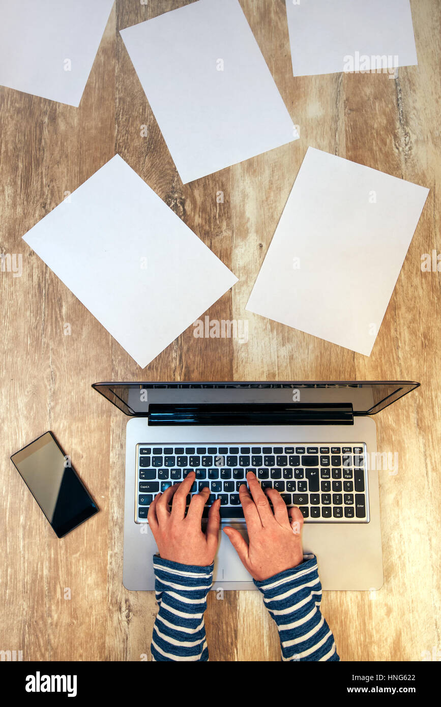 Creative female person working on laptop computer in the office, top view of hands typing keyboard on desk with smartphone and papers scattered around Stock Photo