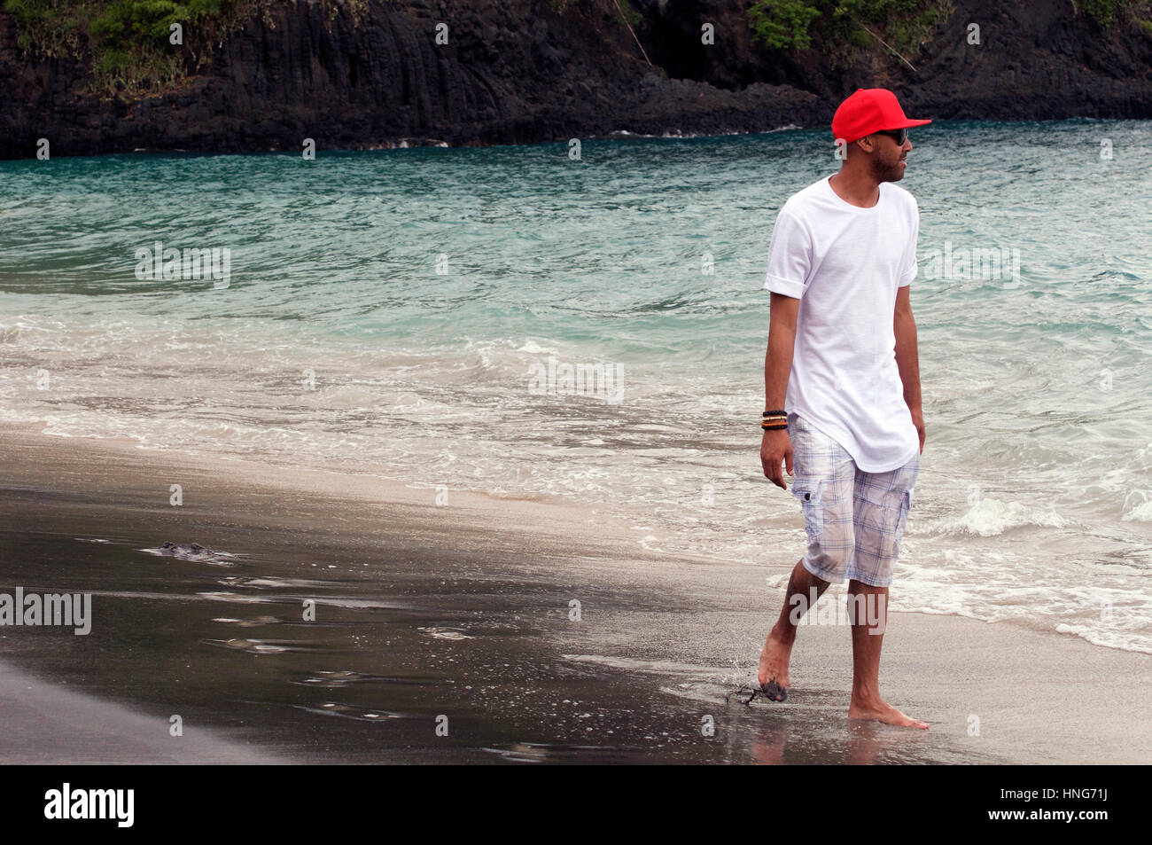 Cool Dude Strolls Along Sandy Beach in Bali Indonesia Wearing Red Baseball Hat, White T-Shirt and Short Pants Stock Photo