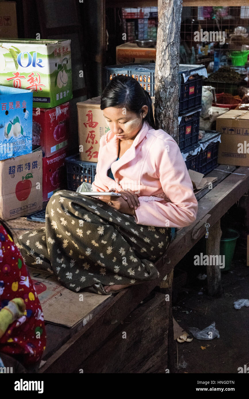 young Burmese girl resting in a Burmese market looking at her Kindle or mobile phone Stock Photo