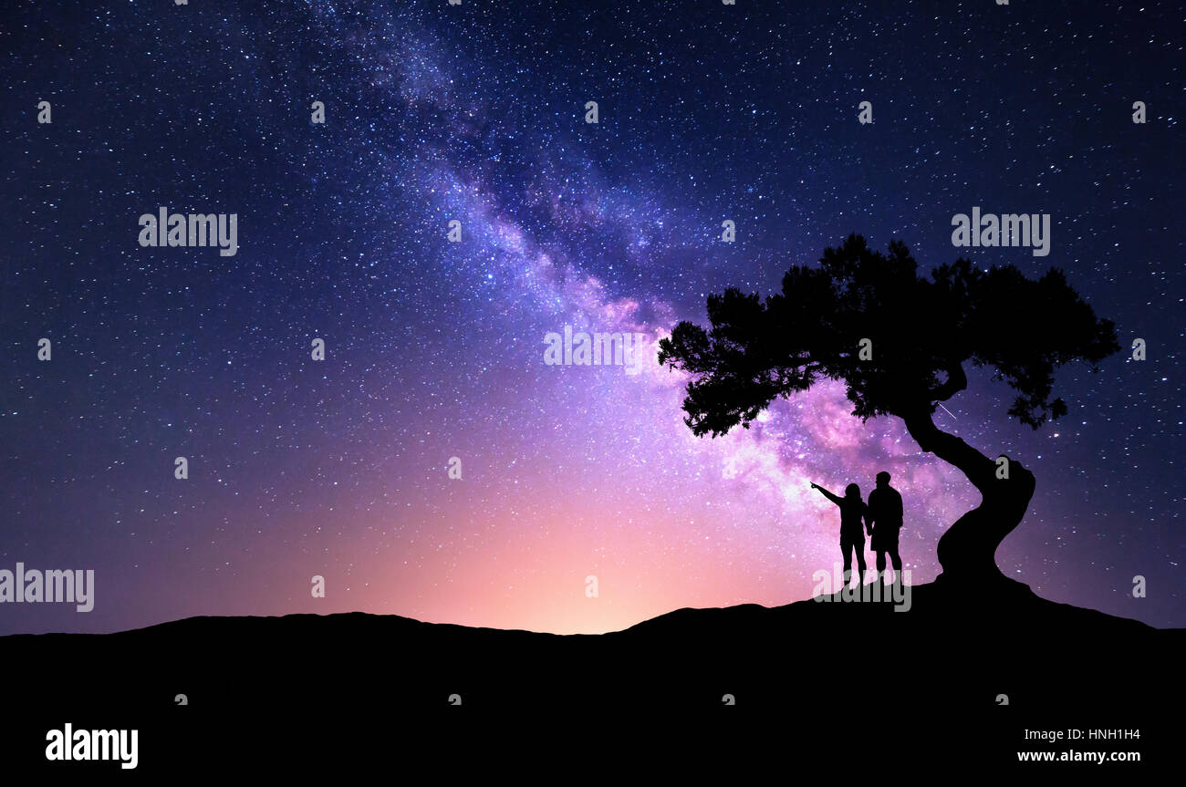 Milky Way with people under the tree on the hill. Landscape with night starry sky and silhouette of standing happy man and woman who pointing finger i Stock Photo