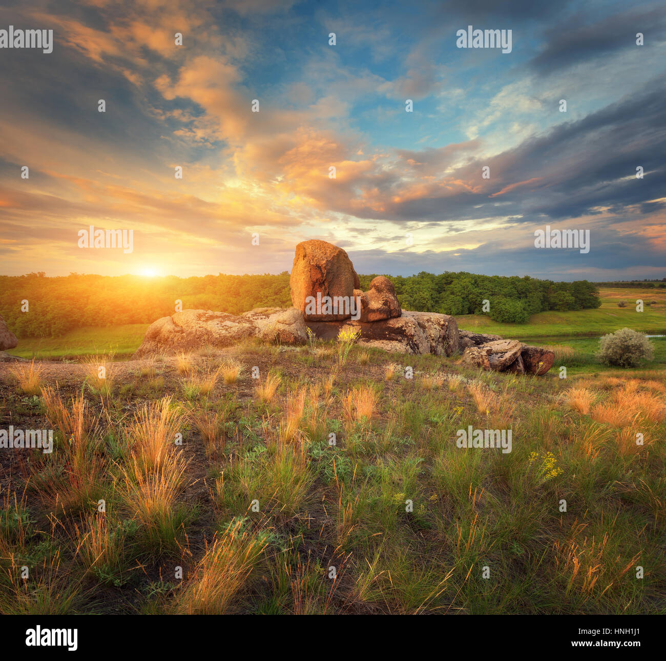 Summer landscape at sunset. Big stones, green and yellow grass against cloudy sky with sunlight. Travel and nature background. Rocks in the beautiful  Stock Photo