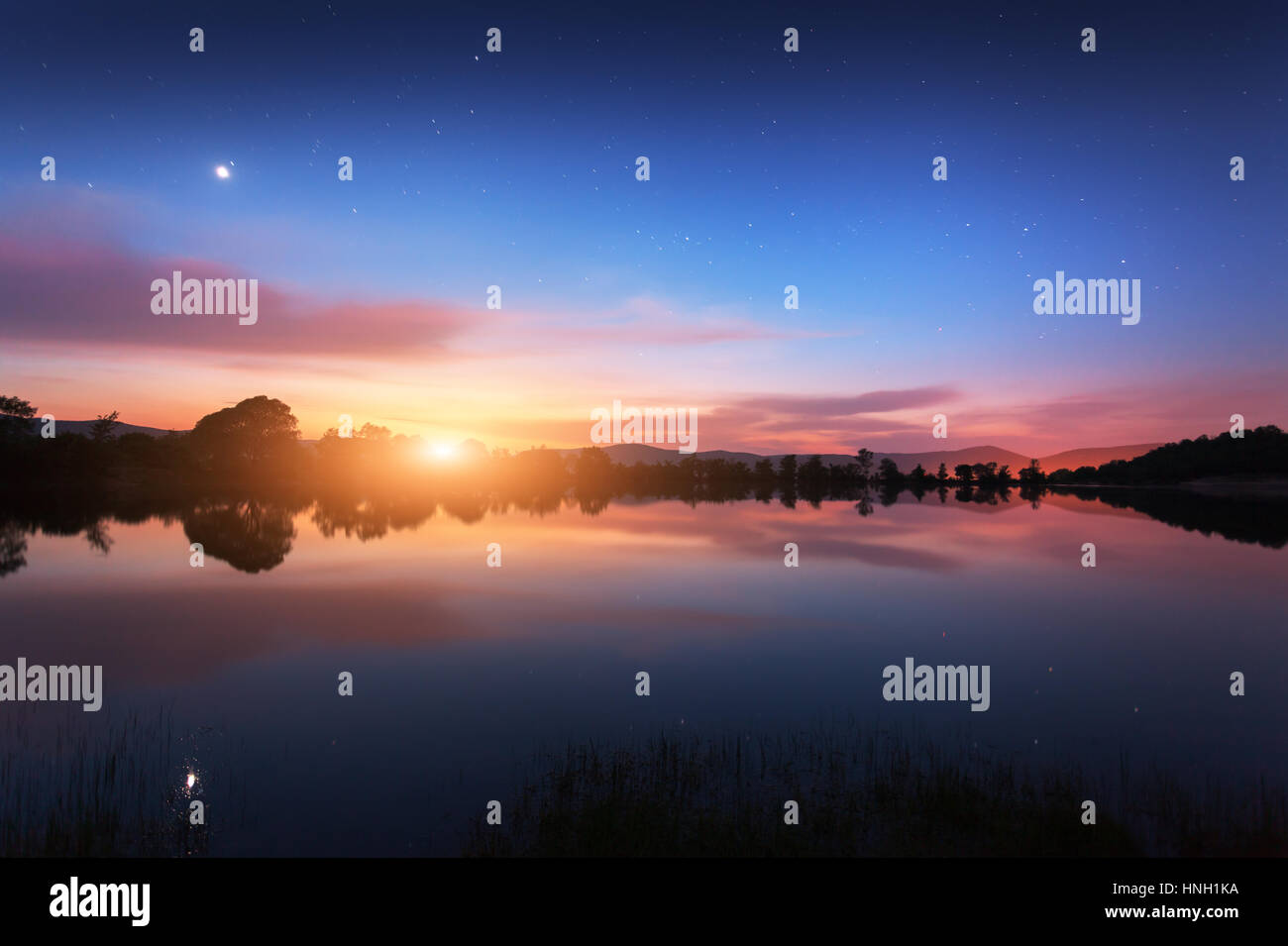 Mountain lake with moonrise at night. Night landscape with river, trees, hills, moon, stars and colorful blue sky with clouds reflected in water. Beau Stock Photo