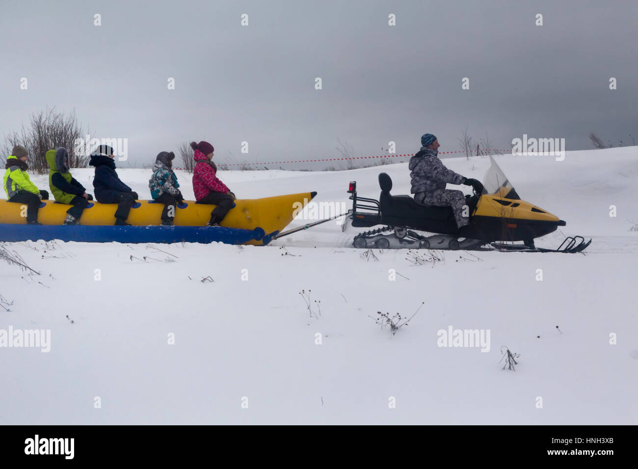 A man on a snowmobile riding children on a banana on a snow-covered valley near Uglich town , Yaroslavl region, Russia Stock Photo