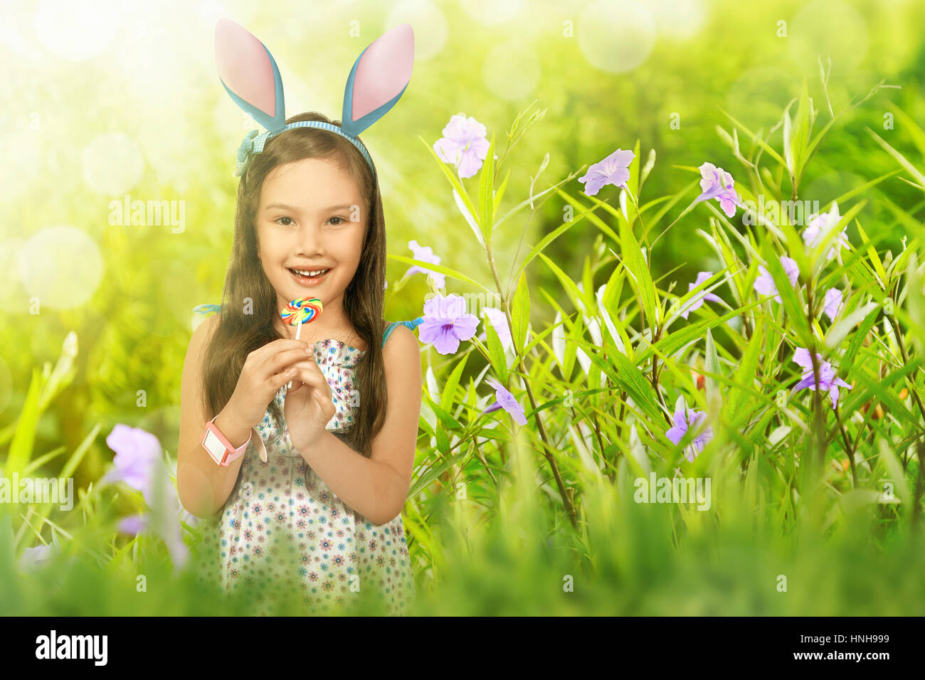 Cute asian little child girl with lollipop wearing bunny ears on green field background Stock Photo