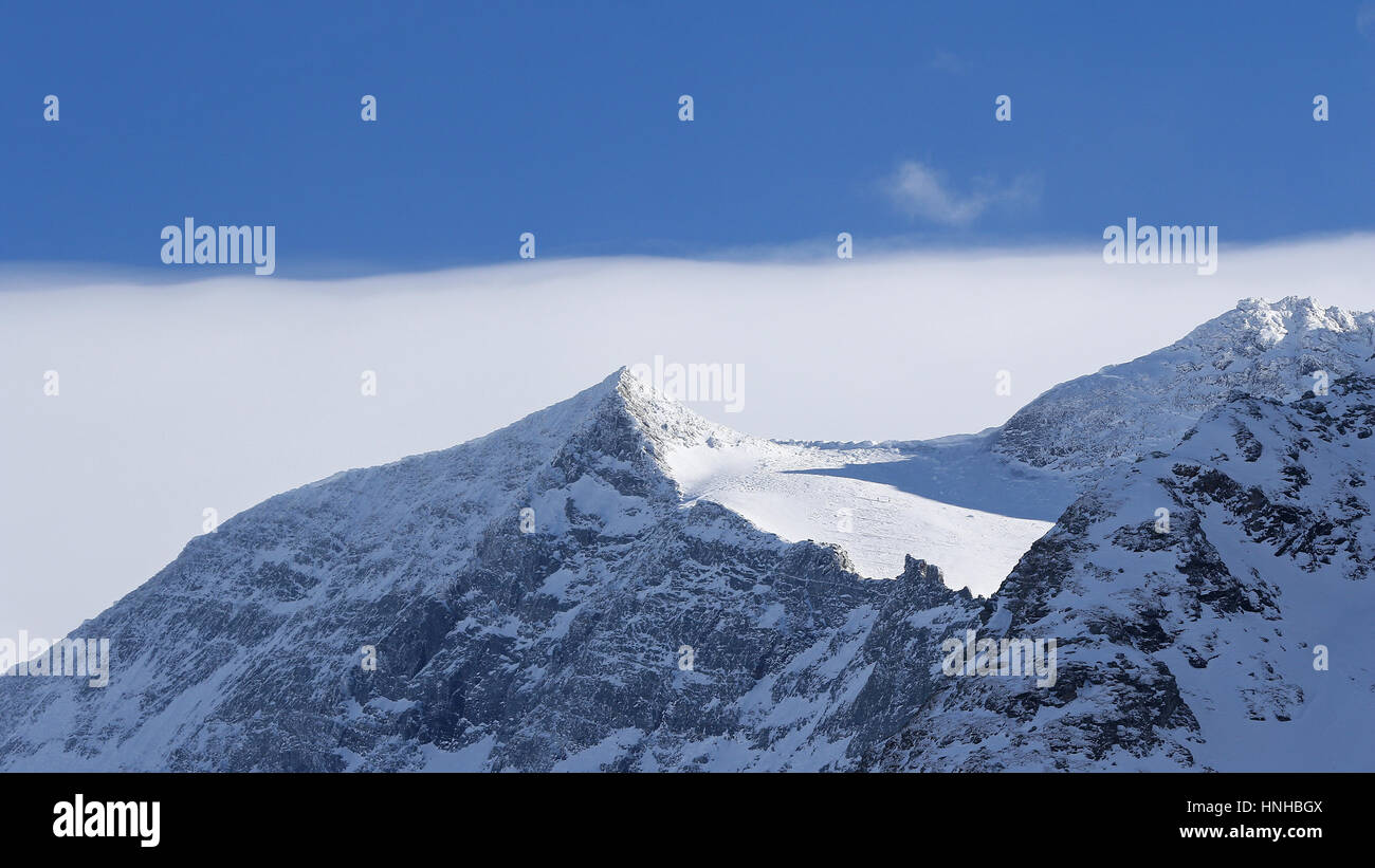 High snow covered mountain peaks in the winter against blue skies with fog Stock Photo