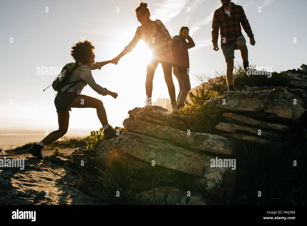 Group of hikers on a mountain. Woman helping her friend to climb a rock. Young people on mountain hike at sunset. Stock Photo