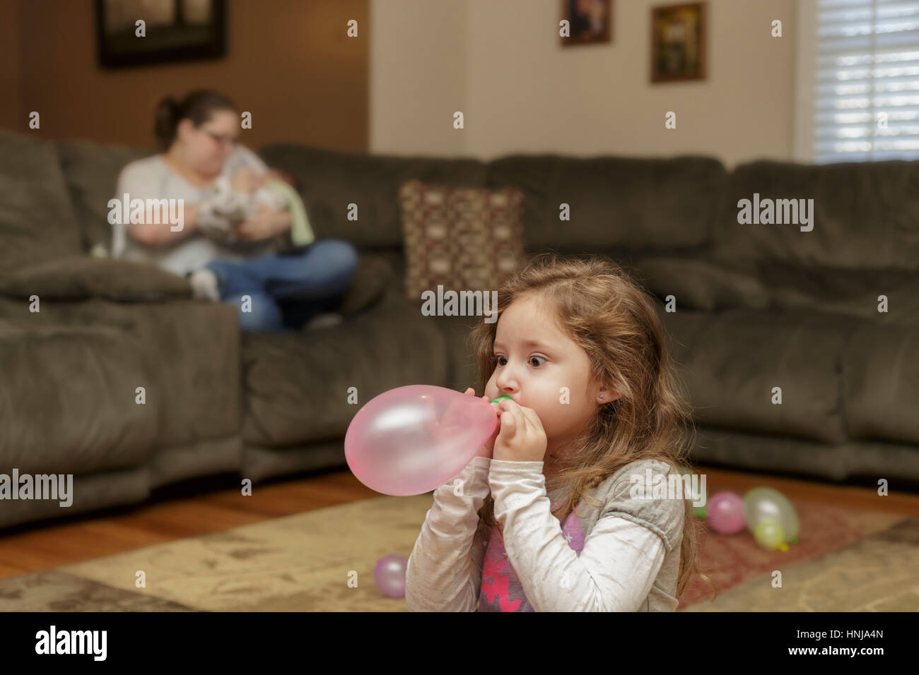 girl blowing up a balloon with mother in the background Stock Photo