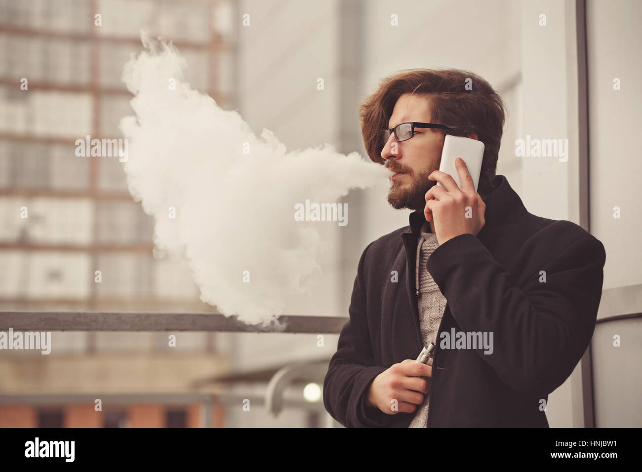 Young hipster man with a beard and glasses smokes an electronic cigarette. He speaks on the cell phone. Youth fashion. Delicious pairs. Cloud of steam Stock Photo