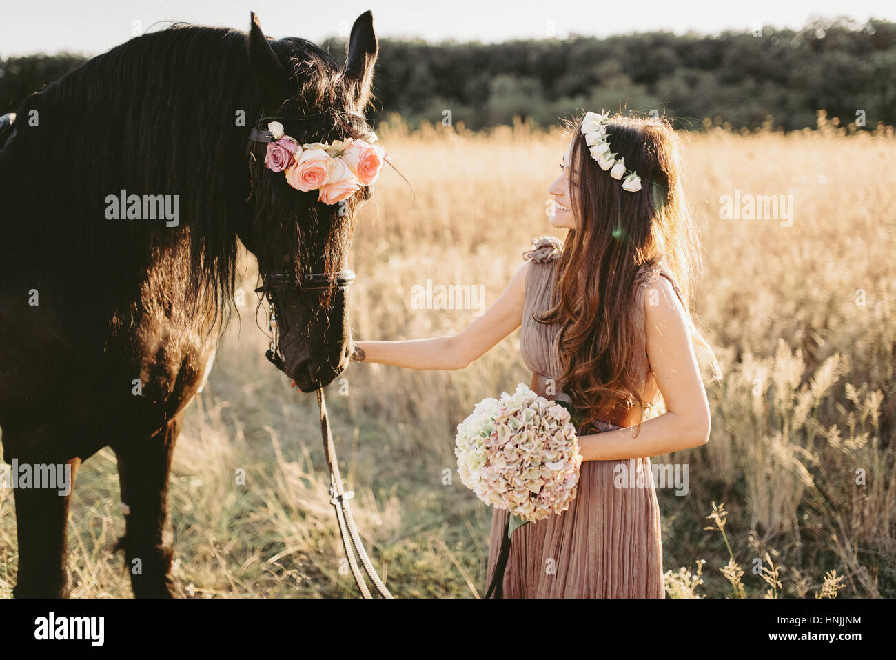 Caucasian female wearing a beige flowy dress and a flower crown, on a yellow grass field with her black horse Stock Photo
