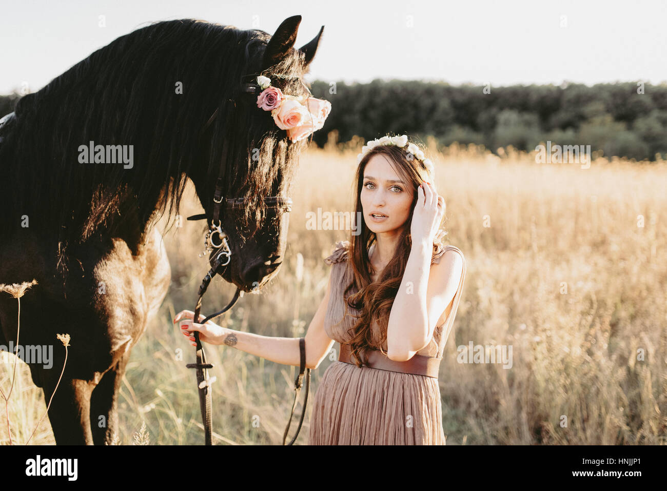 Caucasian woman wearing a beige flowy dress and flower crown, holding the ropes of her black horse, looking at the camera Stock Photo