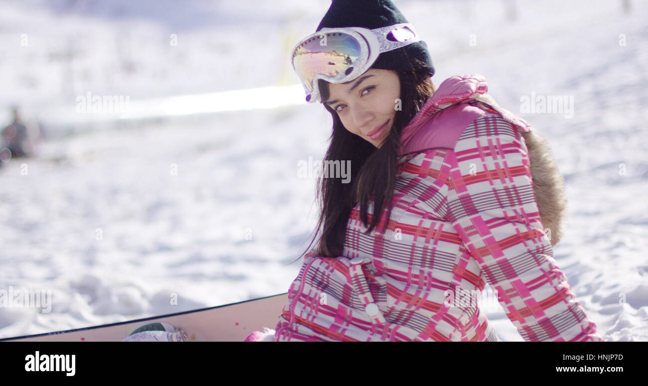 Beautiful asian snowboarder sitting on snow and relaxing. She looking into camera direction with smile. Wearing pink outfit and goggles. Stock Photo