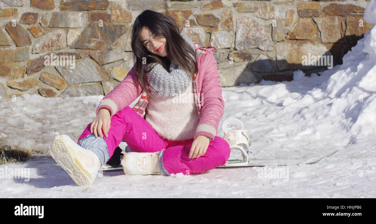 Young asian snowboarder girl relaxing while sitting on snow on her board. She wearing pink ski clothes. With stone wall behind her back. Stock Photo