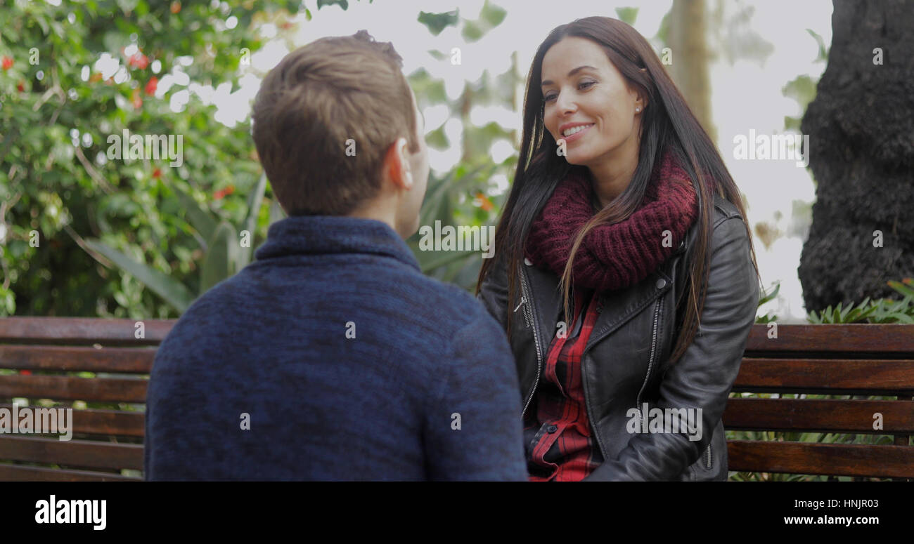 Young man squatting down on the ground talking to his girlfriend as they enjoy a relaxing day in a park sitting on a rustic wooden bench. Stock Photo