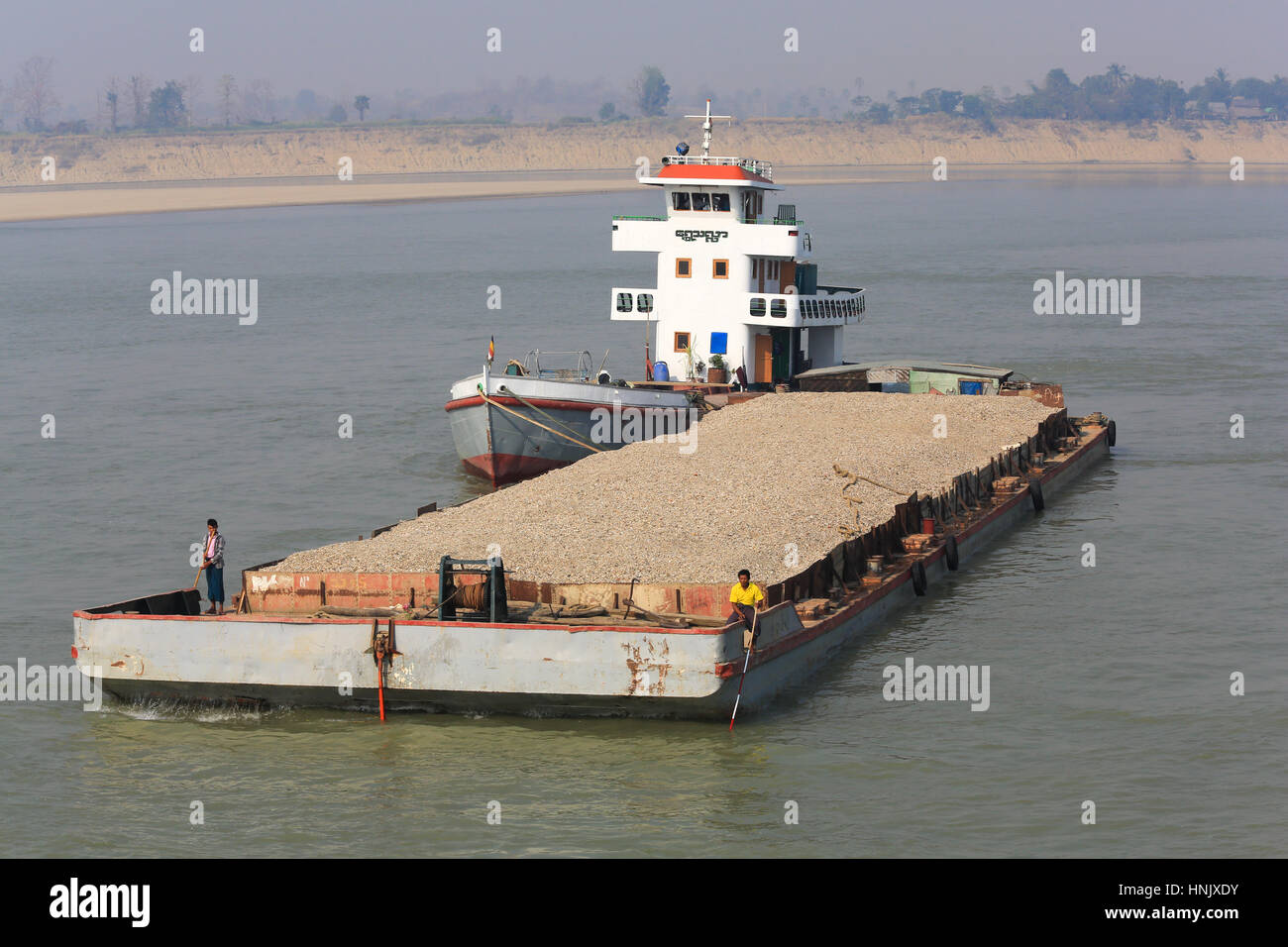 A heavily loaded gravel barge being pushed by a tug boat on the Irrawaddy River while crew use sounding poles to check for adequate depth in Stock Photo