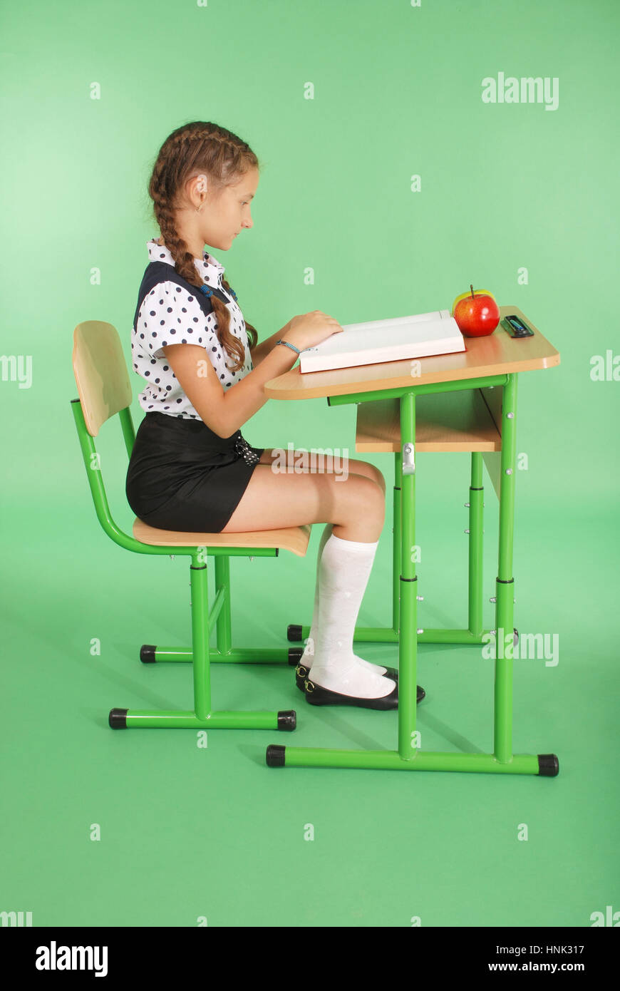 Girl in a school uniform sitting at a desk and reading a book isolated on green Stock Photo