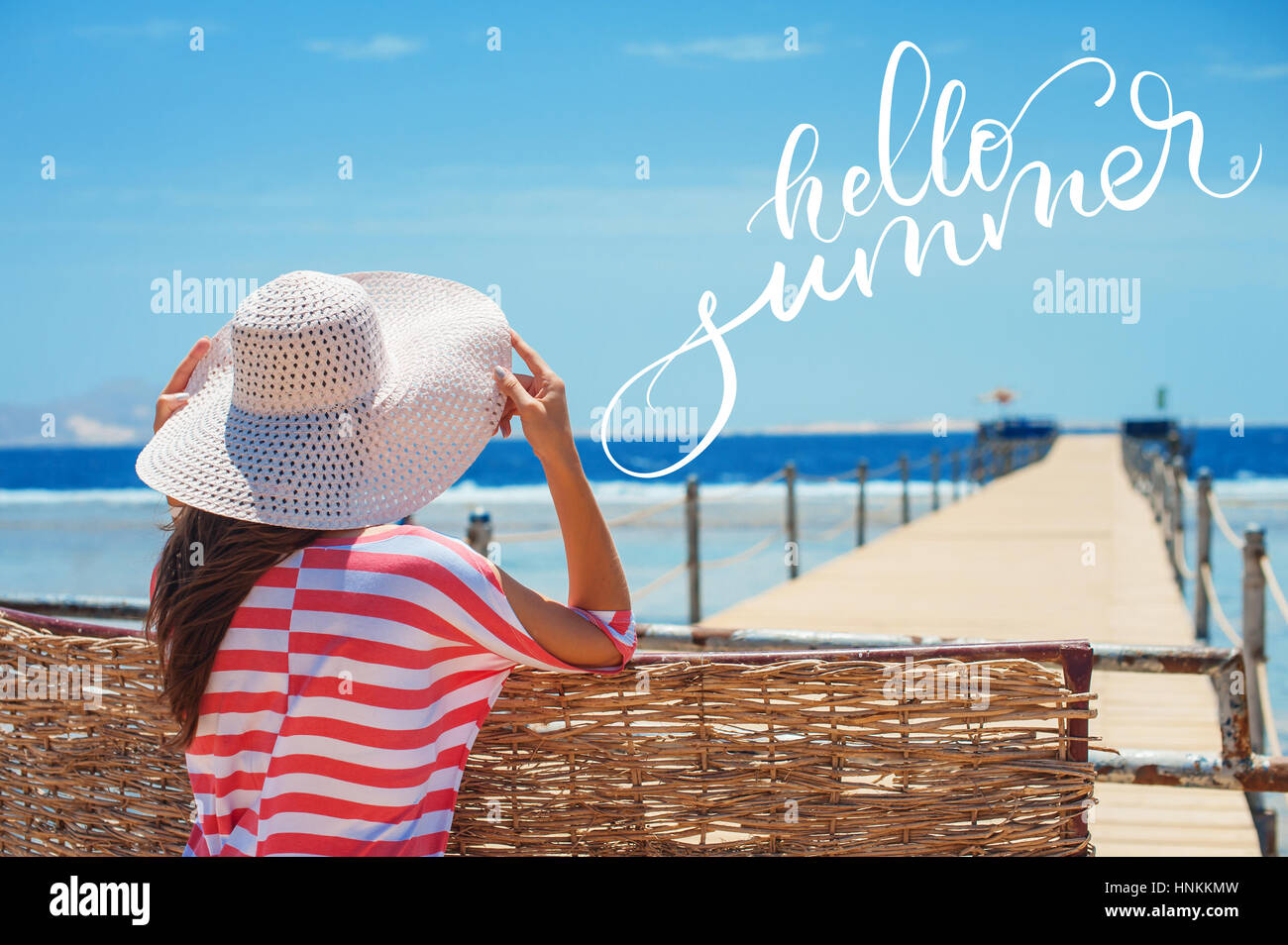 Closeup back view of woman in white hat looking out towards blue ocean and sky and words Hello summer. Calligraphy, lettering Stock Photo