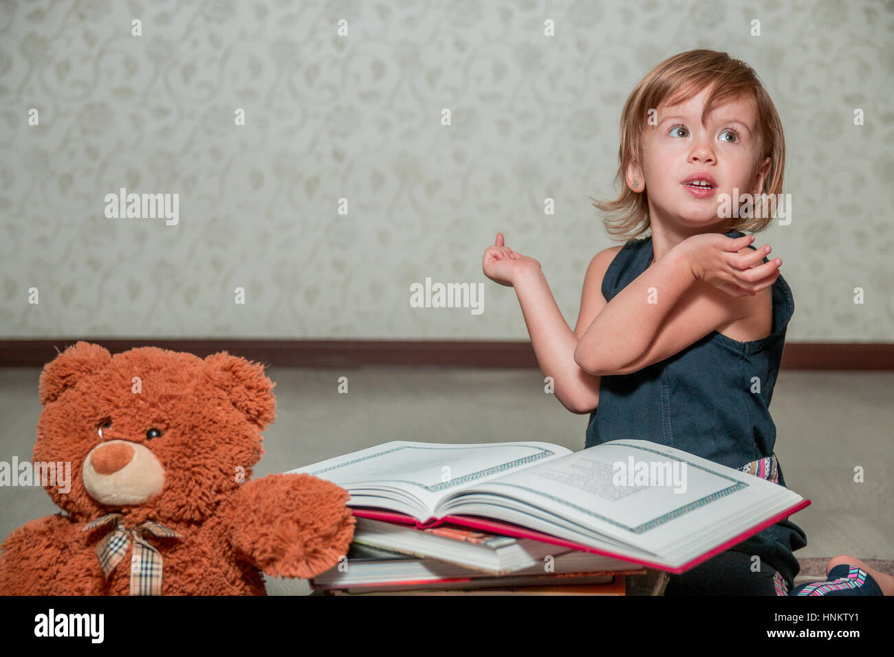 A little girl in a dark blue dress reading a book sitting on the floor ...