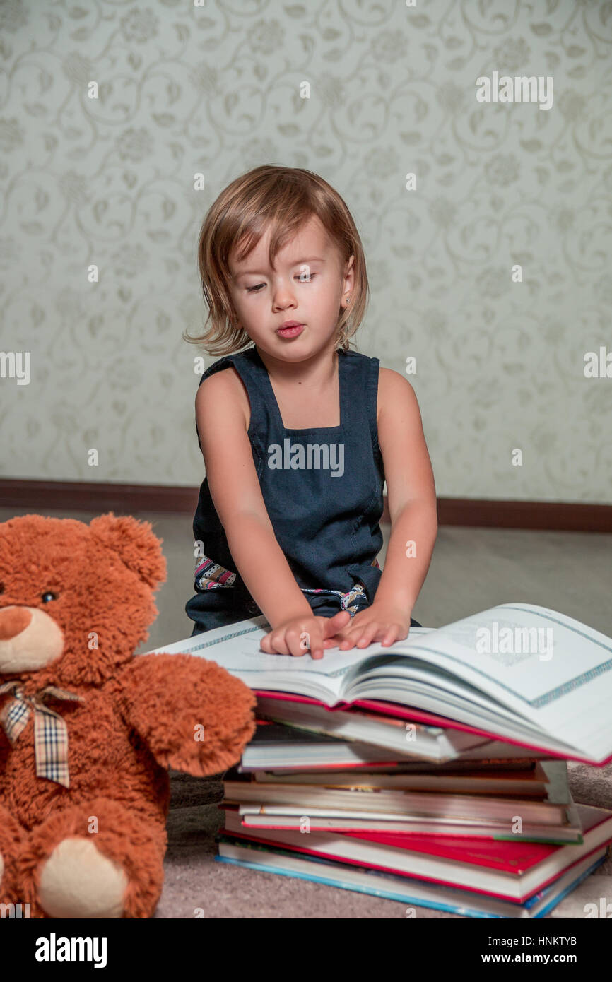 A little girl in a dark blue dress reading a book sitting on the floor ...