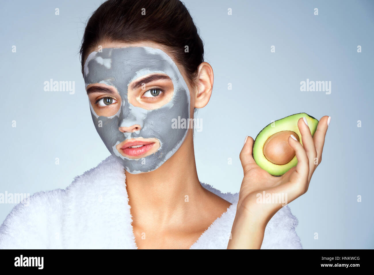 Attractive girl holding half an avocado in hand. Photo young woman with mask of clay on blue background. Beauty & Skin care concept Stock Photo