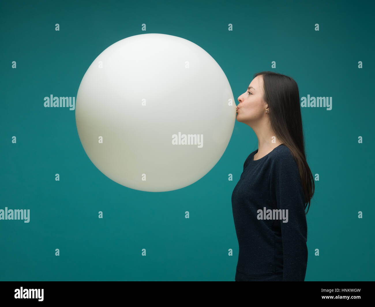 funny young woman with long dark hair inflating huge balloon on blue background Stock Photo