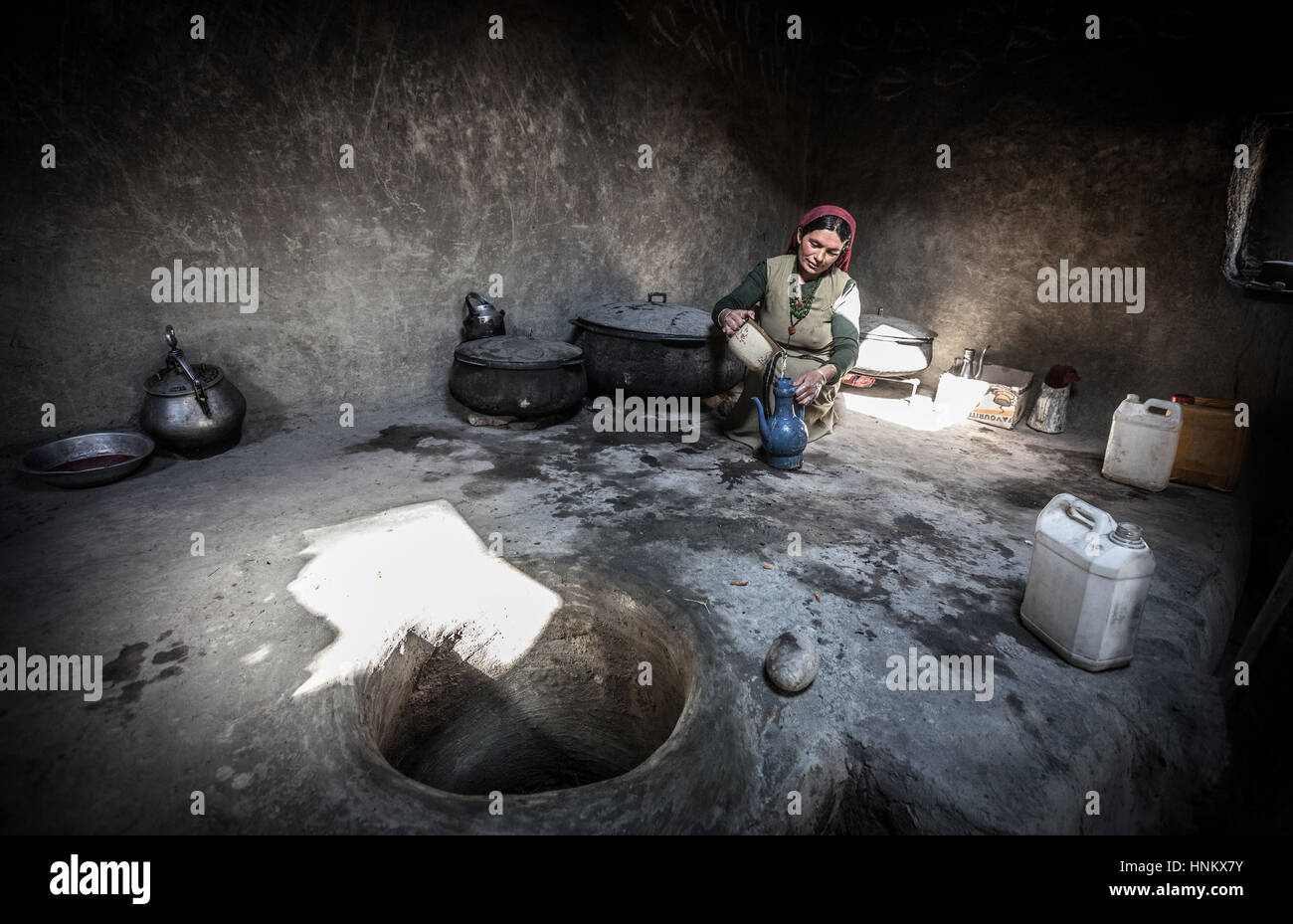 Afghanistan, Wakhan corridor,a woman is pouring water in traditional clothes, seated on the ground in stoned kitchen. Stock Photo