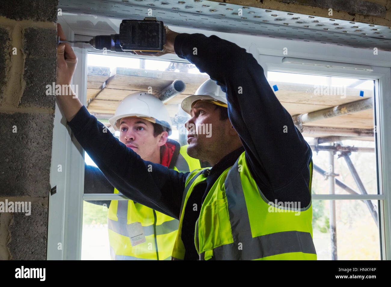 Two workmen on a construction site, builder in hard hat using an electric drill on a window frame. Stock Photo